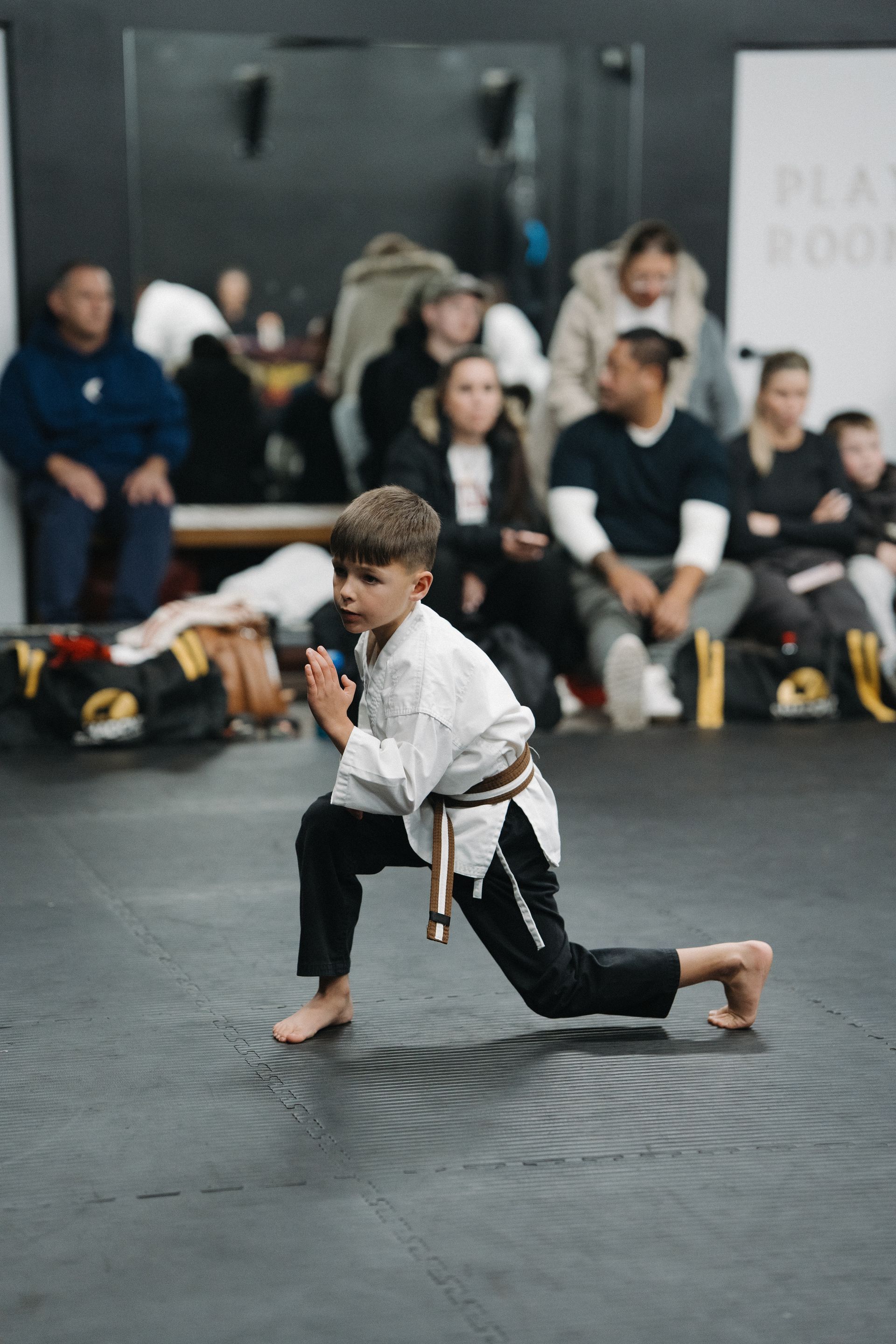 Boy in karate uniform performing a low lunge with onlookers in the background.