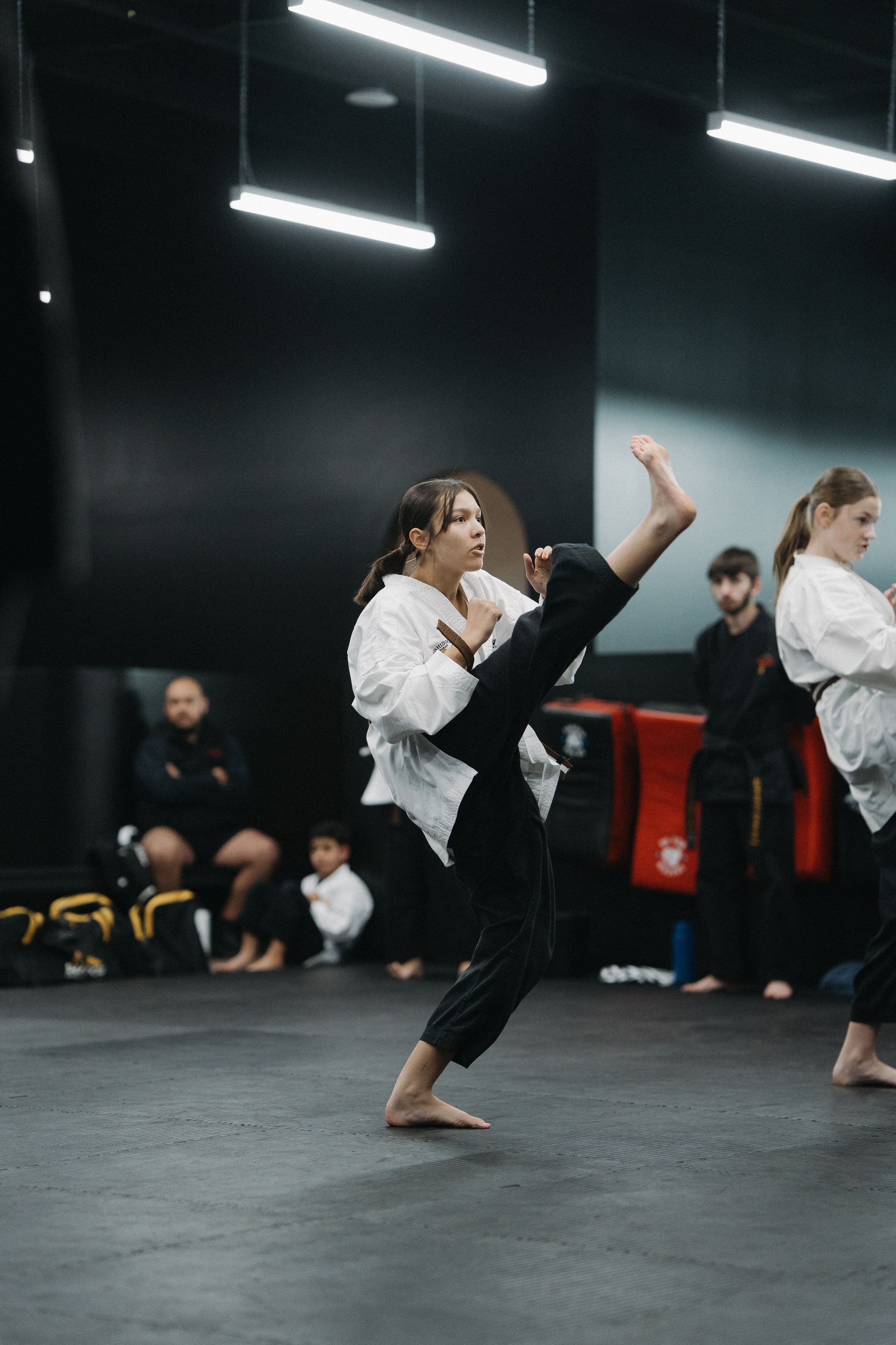 Person in karate uniform performs a high kick in a dojo. Others observe.