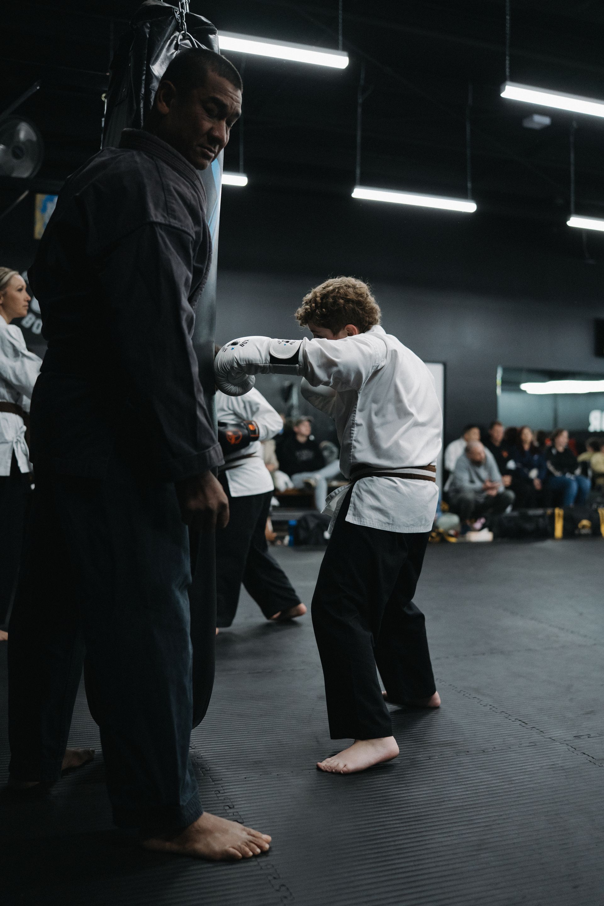 Person in black uniform holds a punching bag as another practices a punch in a martial arts studio.