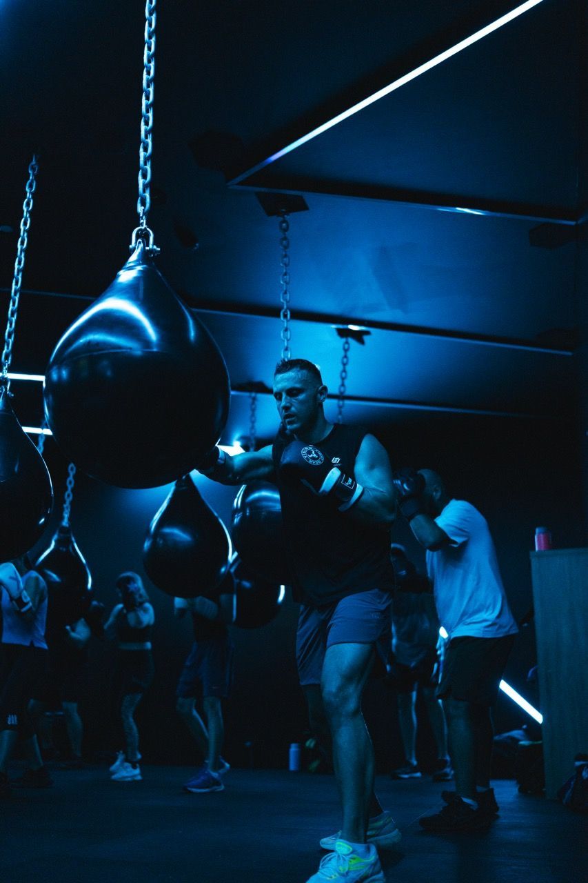Man boxing a teardrop bag in a dimly lit, blue-hued gym. Others are also working out.