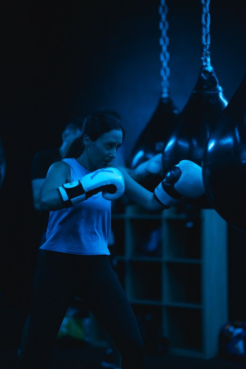 Woman boxing, wearing gloves, in a blue-lit gym, punching a bag.