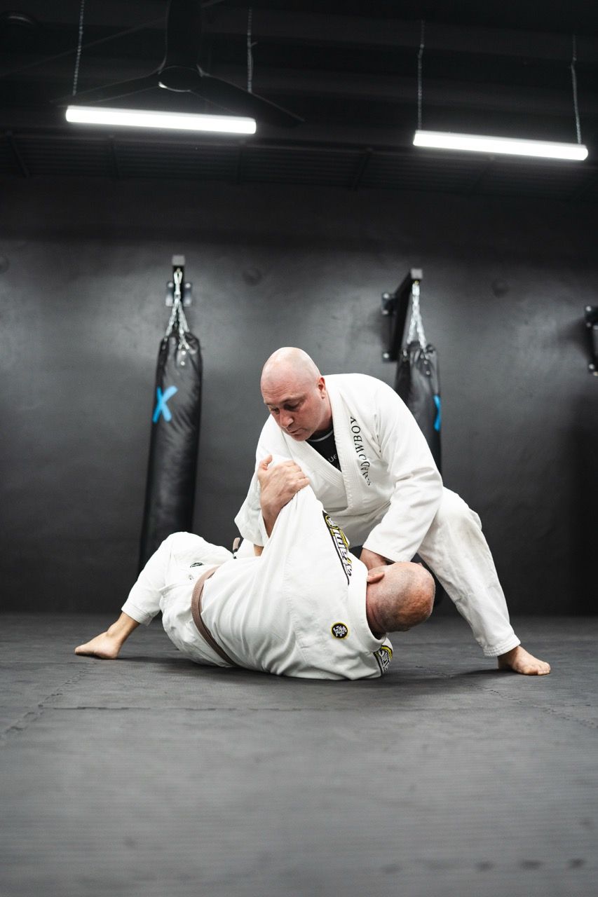Two men in white martial arts uniforms practice on a gym mat; one is on top in a dominant position.