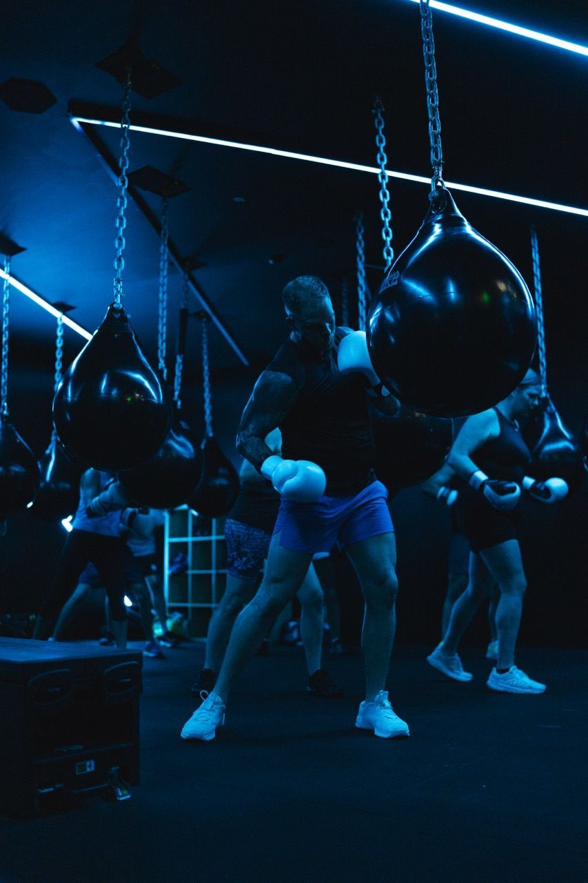 People boxing in a dark gym, striking teardrop-shaped bags. Blue lighting and chains visible.