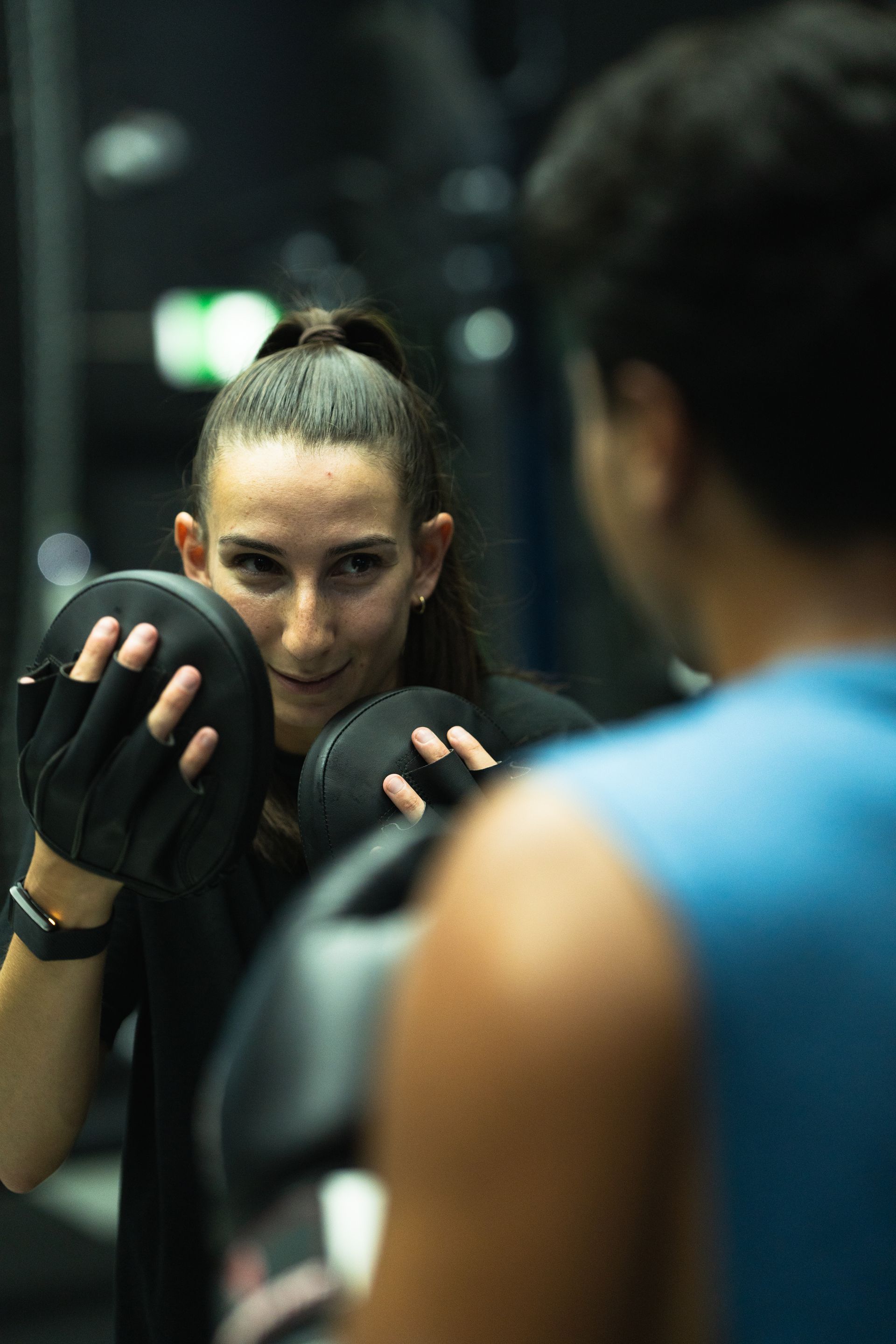 Woman holding mitts, facing a person in a gym setting, smiling; the person is wearing a boxing glove.
