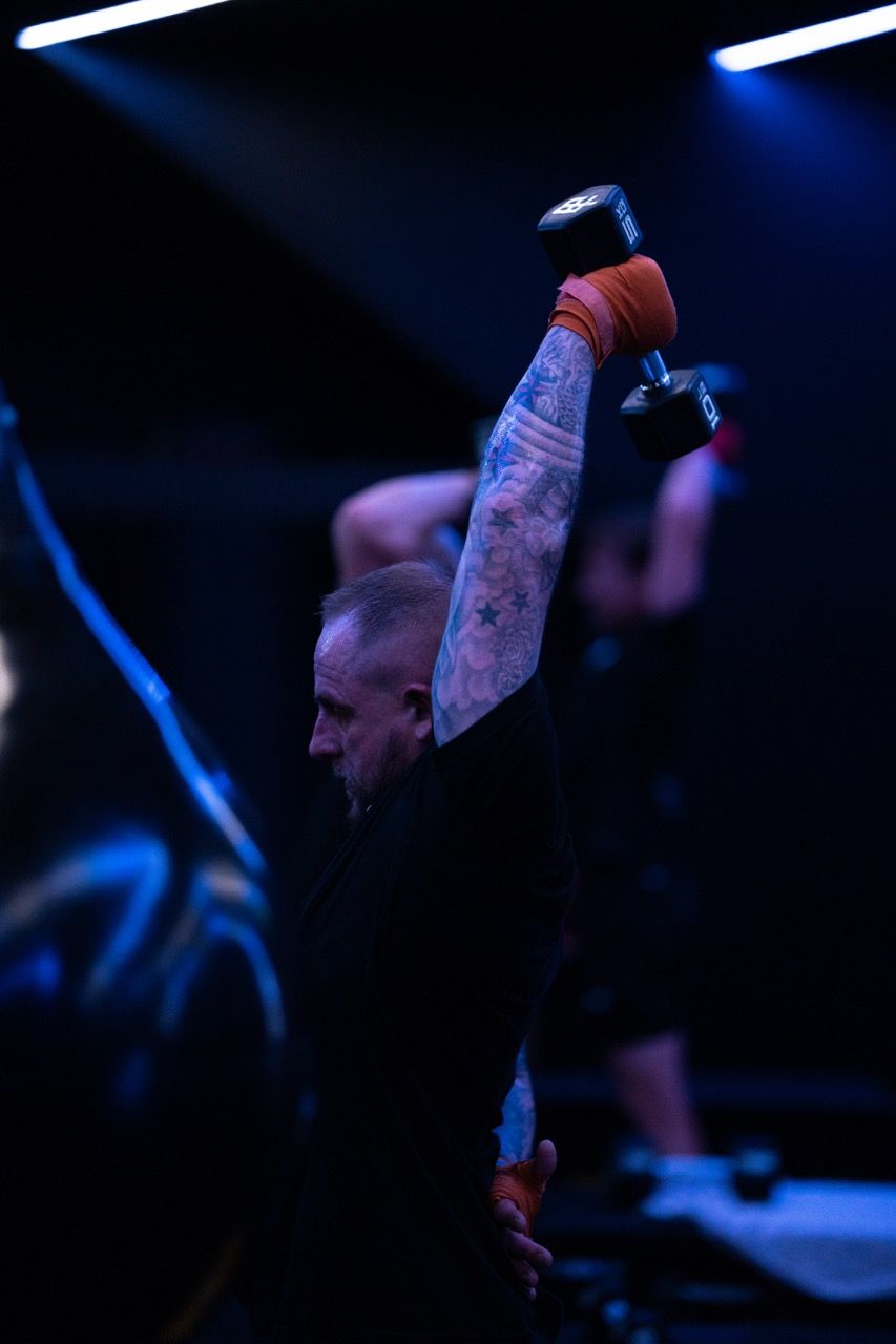 Man with arm tattoos lifting a dumbbell overhead in a dark gym, other person in background also lifting.
