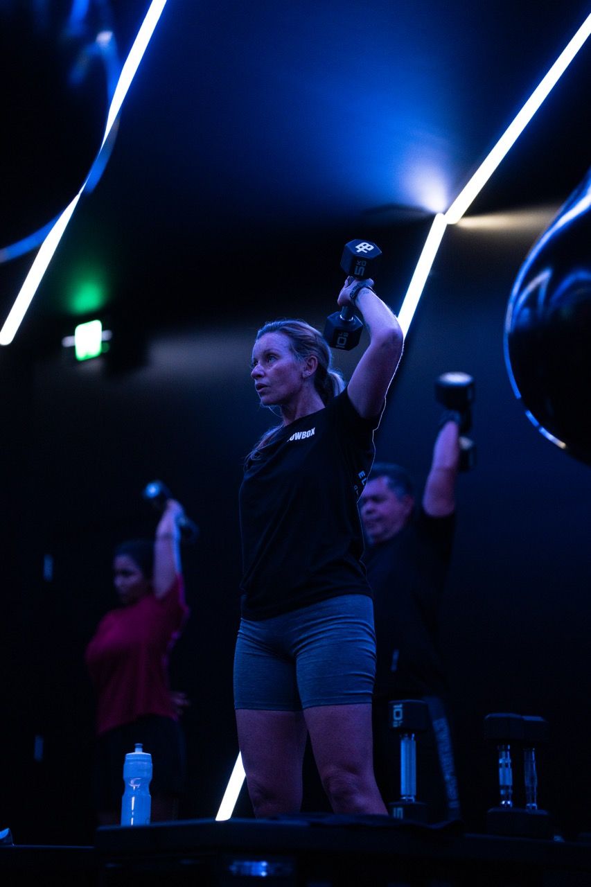 People in a gym exercising with dumbbells. Blue-lit room. Woman in center lifts weight above head.