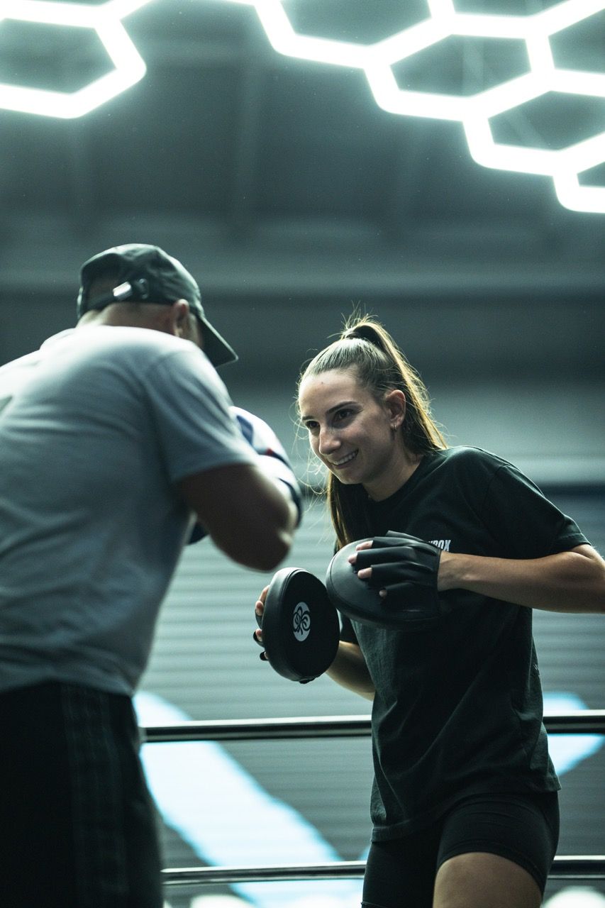 Woman boxing with a trainer in a ring, lit by honeycomb-shaped lights. Both wear gloves; she smiles.