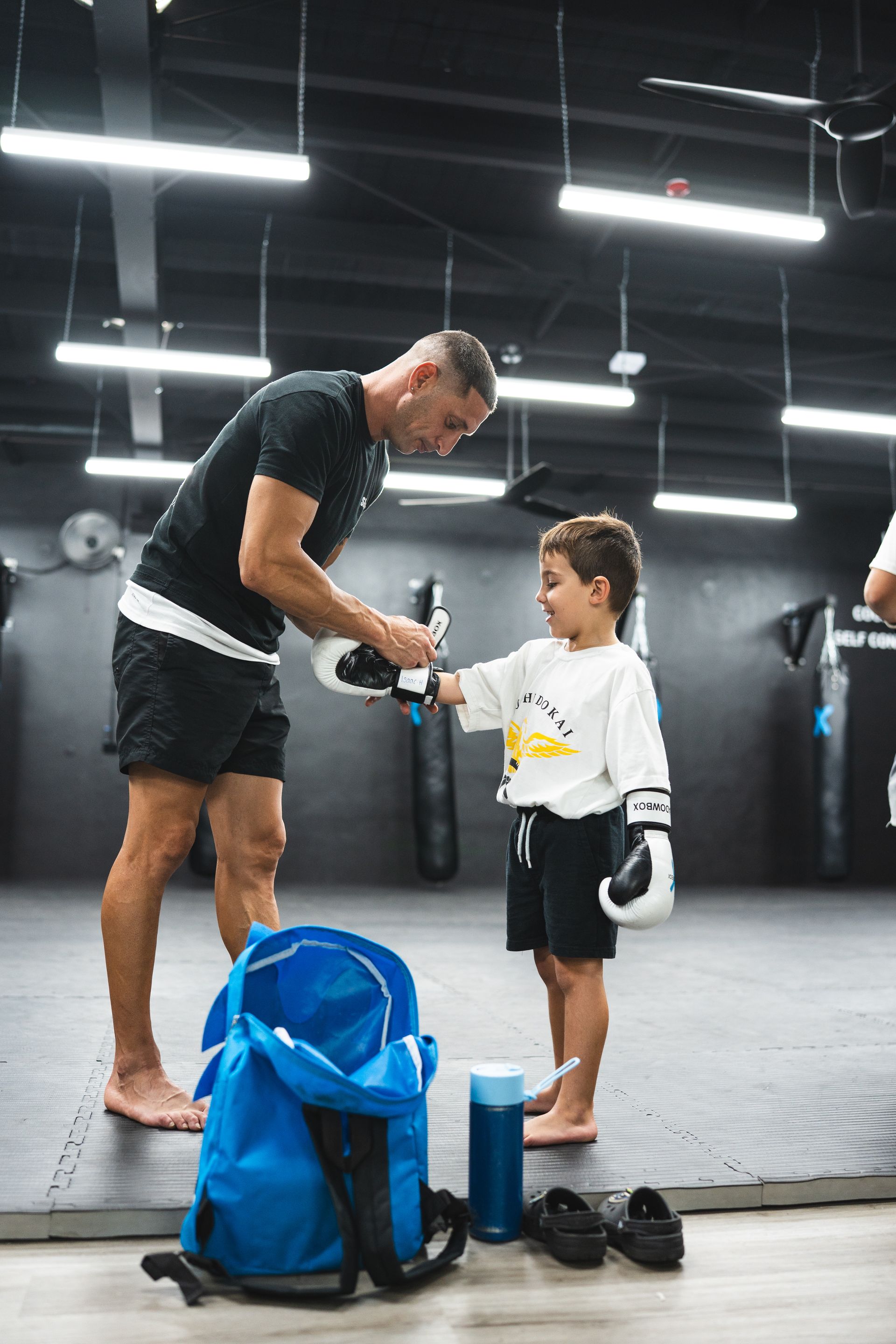 A man fitting boxing gloves on a young child in a gym, blue bag, black shoes nearby.
