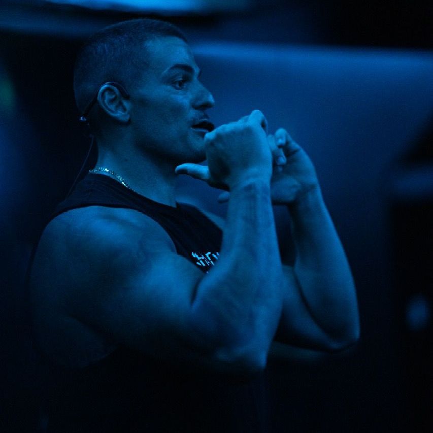 Man in a black tank top gestures with hands, illuminated by blue light.