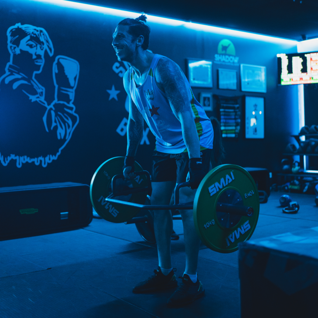 Man lifting weight bar in a gym with blue lighting; smiling.