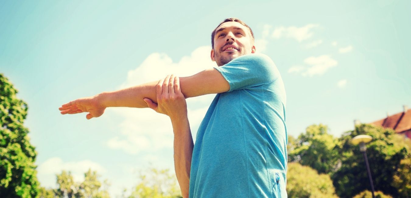 Man stretching his arm outside, blue shirt, sunny day.