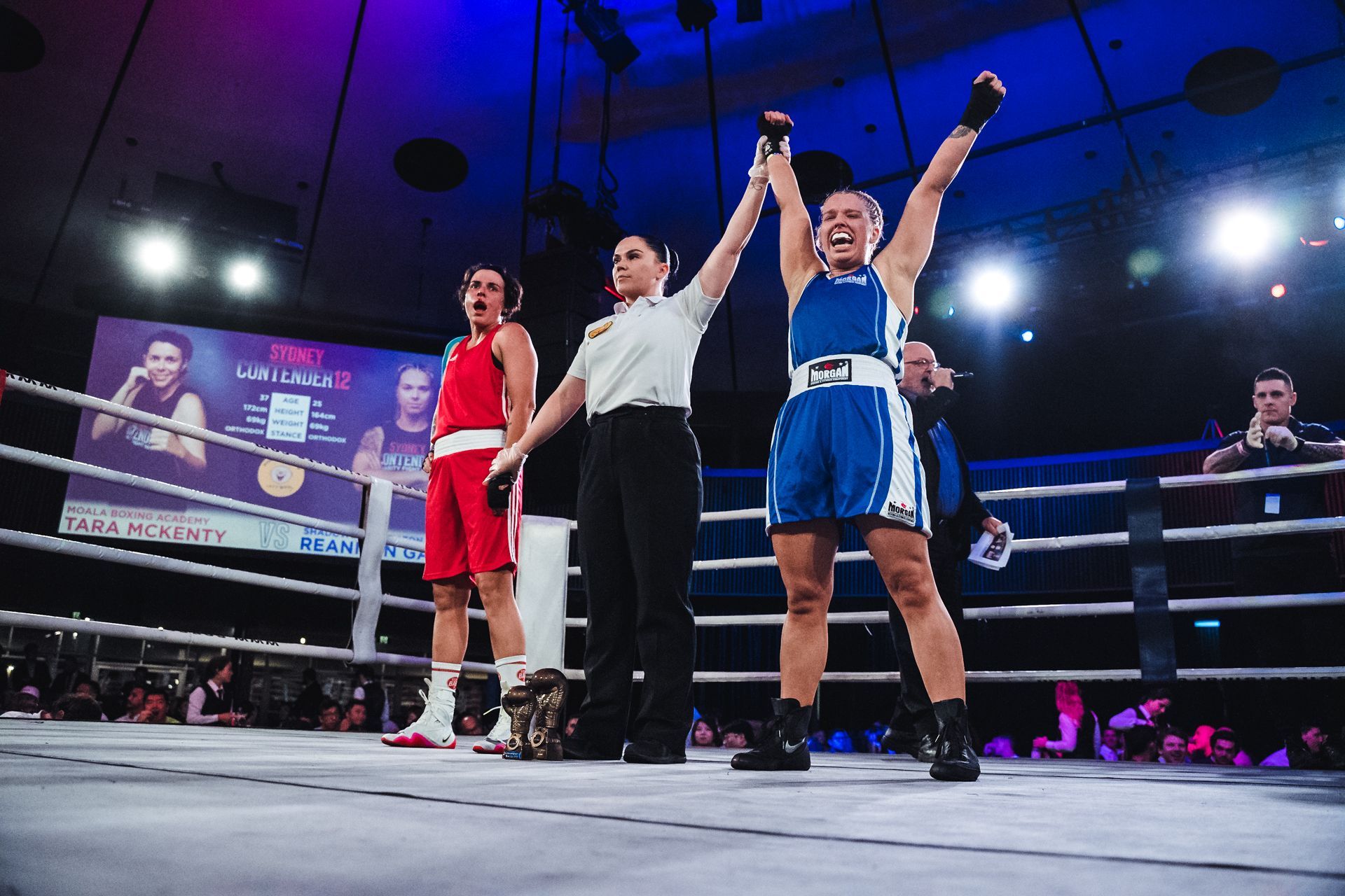 Boxer in blue raises arms victoriously in boxing ring, referee between her and opponent in red.
