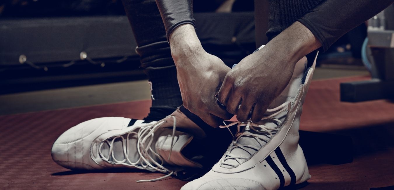 Person tying the laces of white boxing boots in a gym setting.