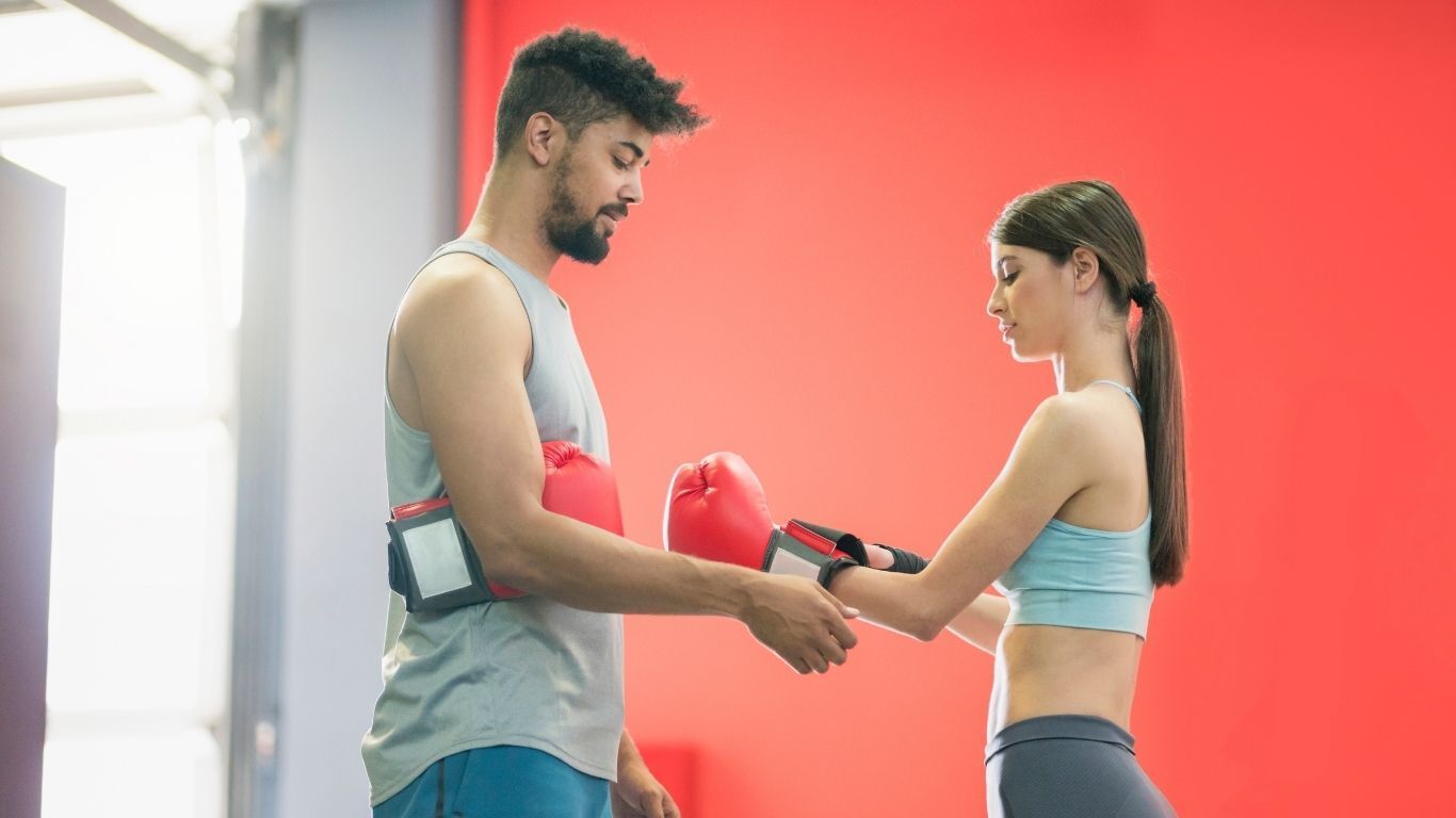 A man in a tank top helps a woman in a sports bra put on boxing gloves in a gym. Red background.