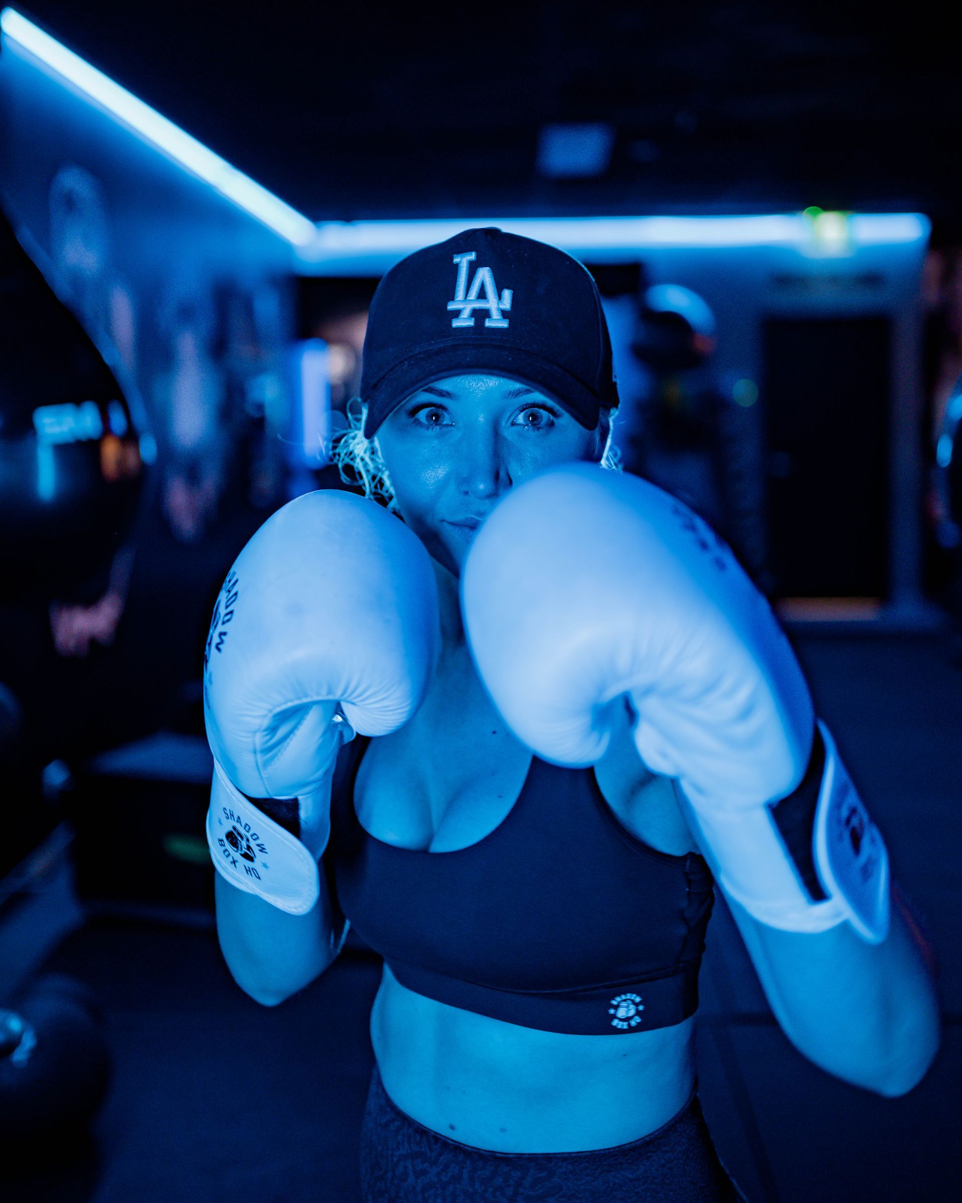Woman in boxing gloves, cap, and sports bra, in a dimly lit, blue-toned boxing gym, in fighting stance.