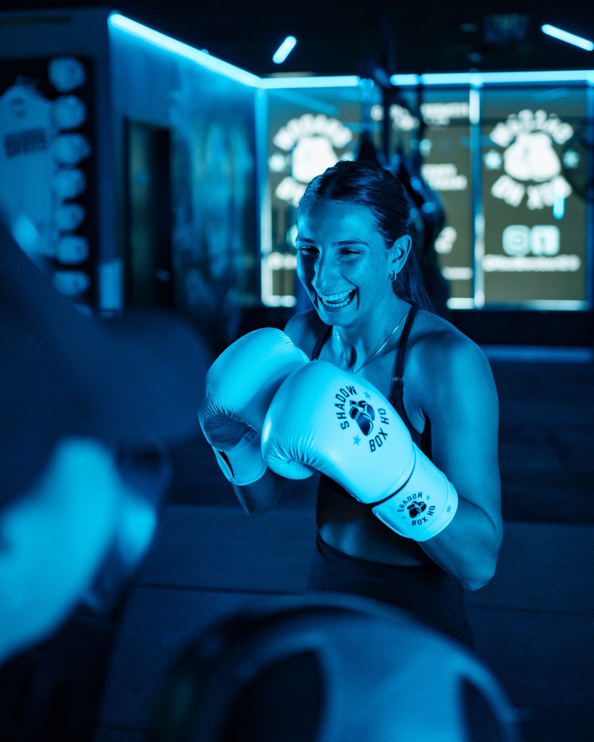 Woman in boxing gloves smiles, working out in a blue-lit gym.