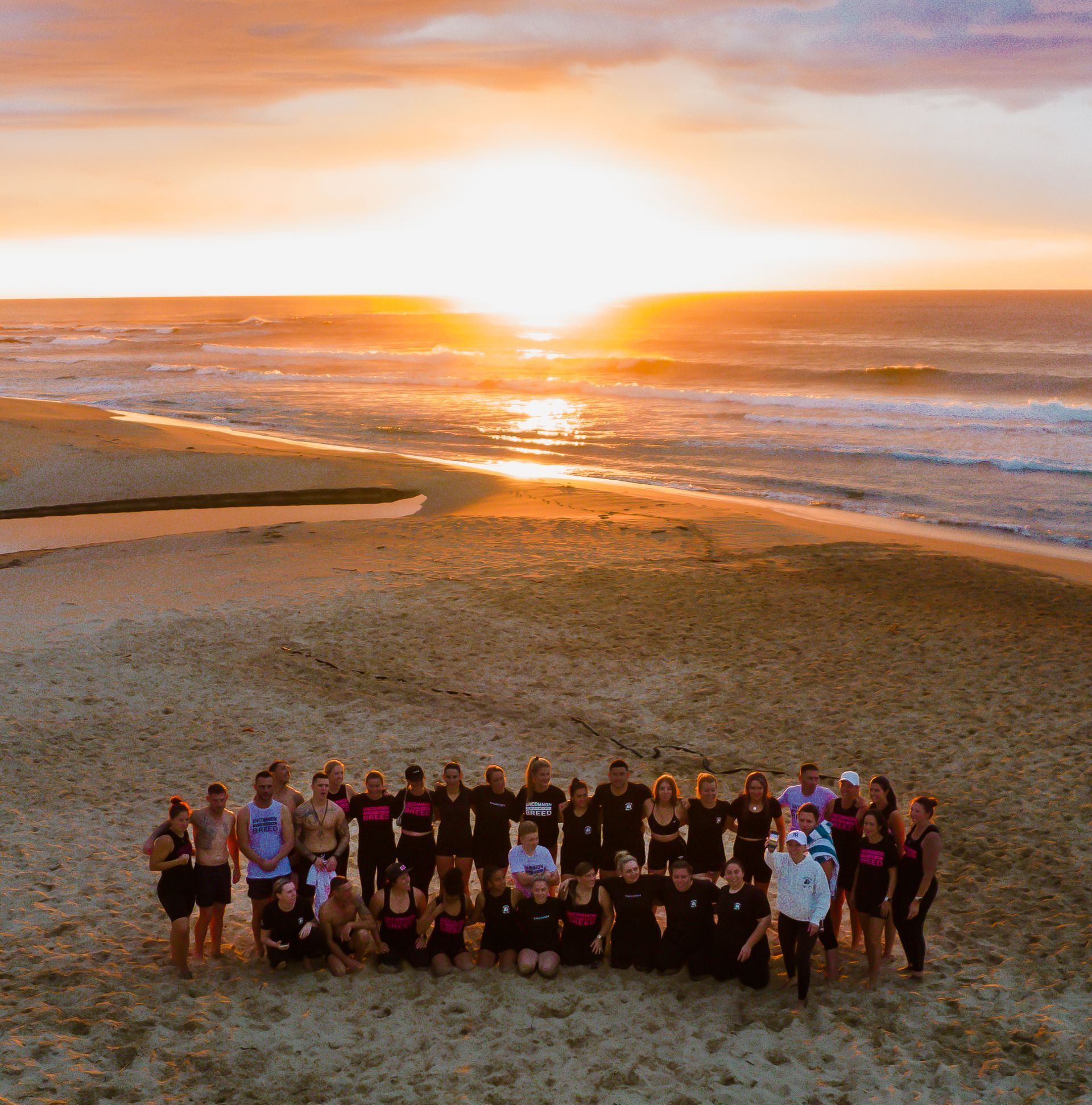 People on beach at sunset. Ocean waves, golden sky, sand. Group in dark and pink shirts.