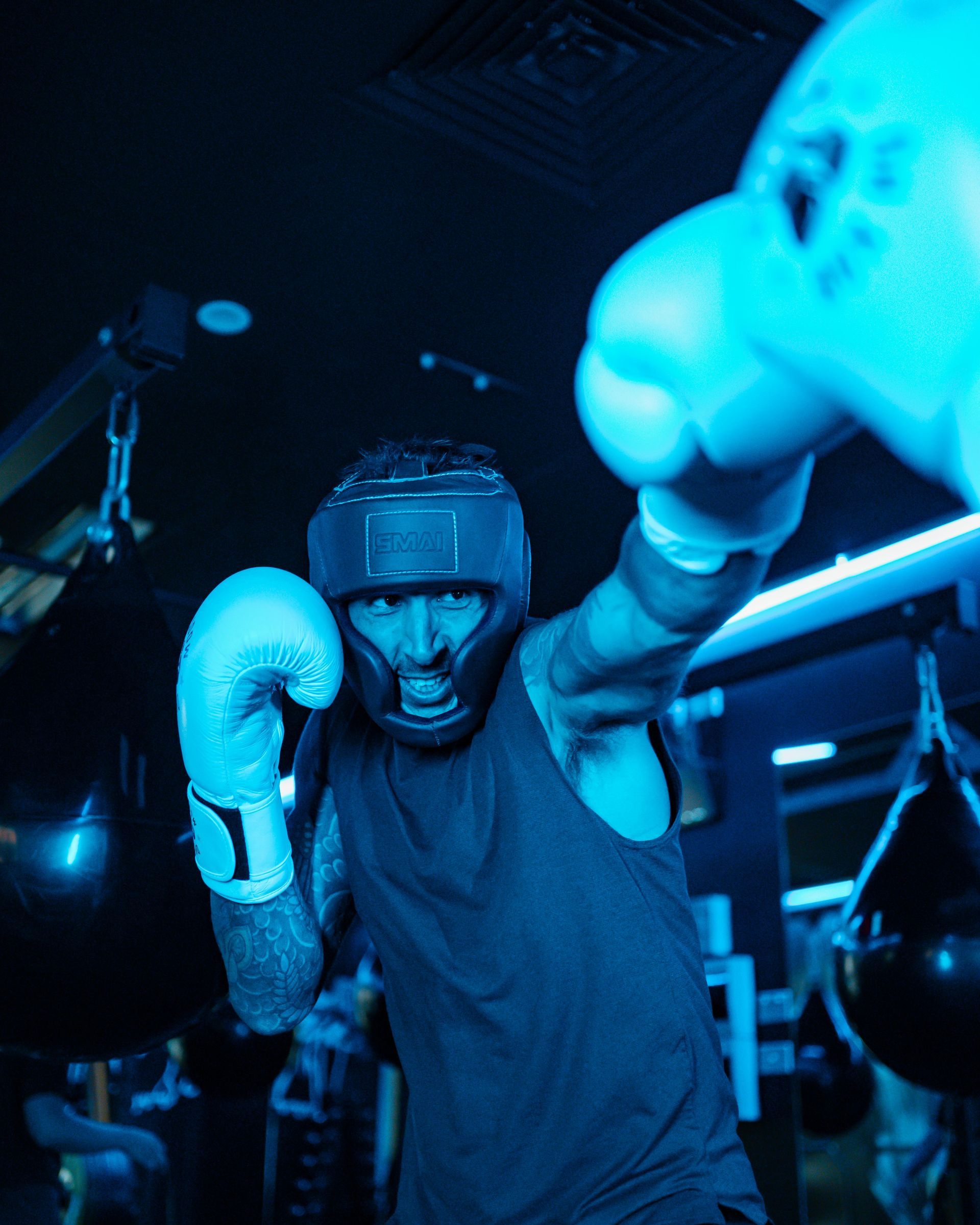 Boxer in protective gear throwing a punch inside a gym, illuminated by blue light.