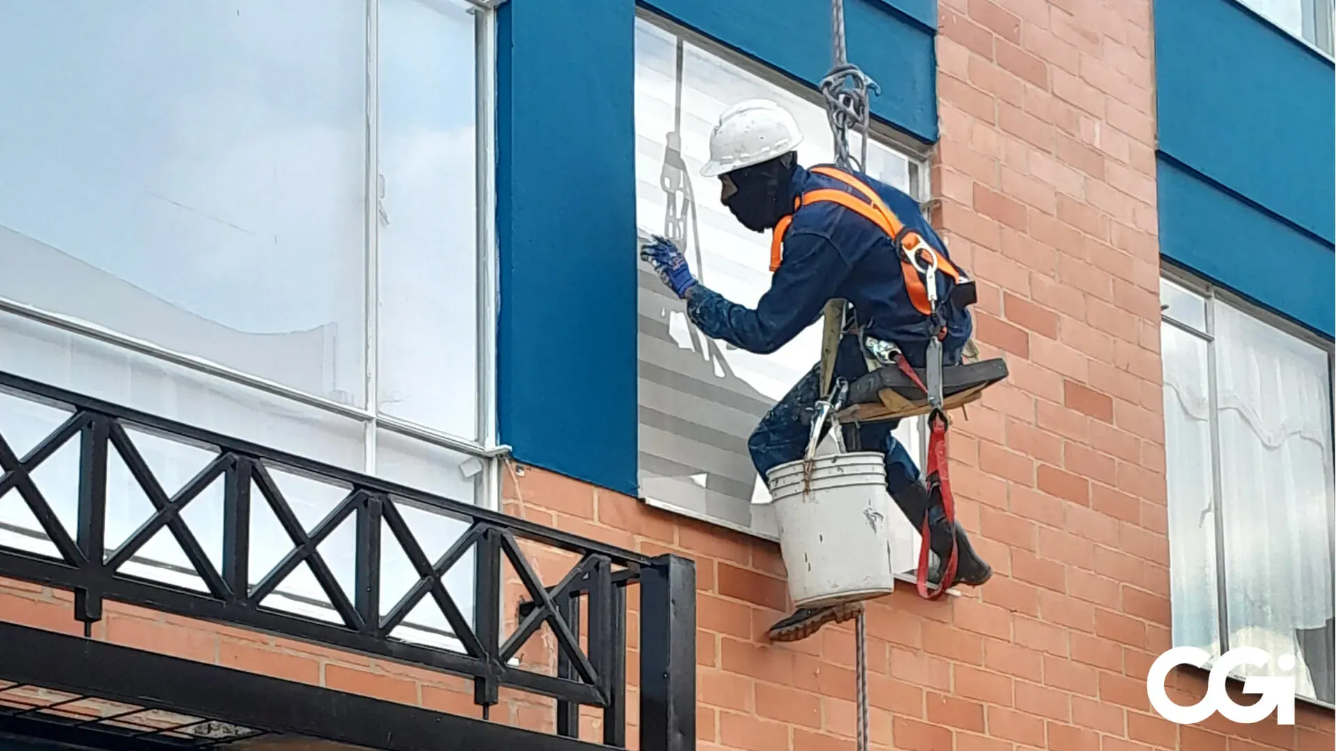 Limpiador de ventanas con equipo de seguridad pintando una moldura azul en un edificio de ladrillo.