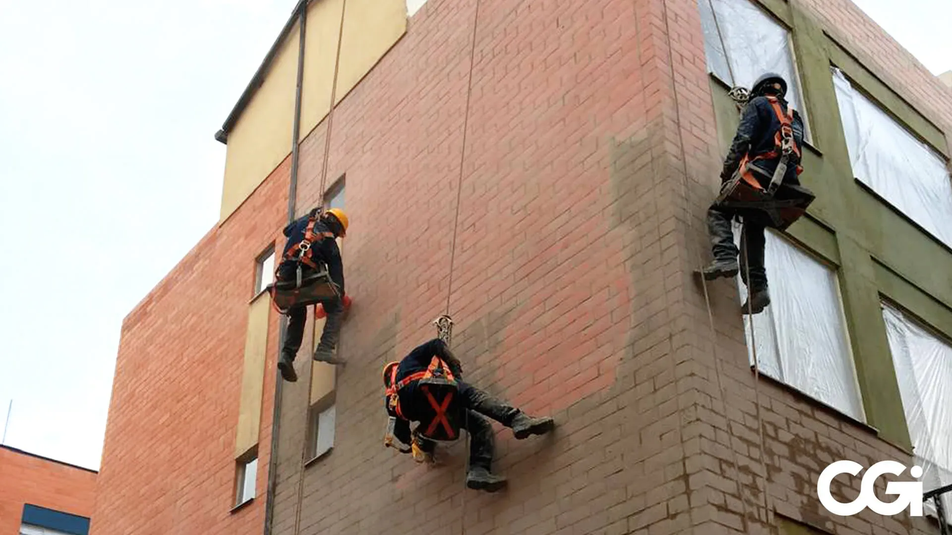 Tres limpiadores de ventanas suspendidos de cuerdas, limpiando un edificio de ladrillos de color naranja.