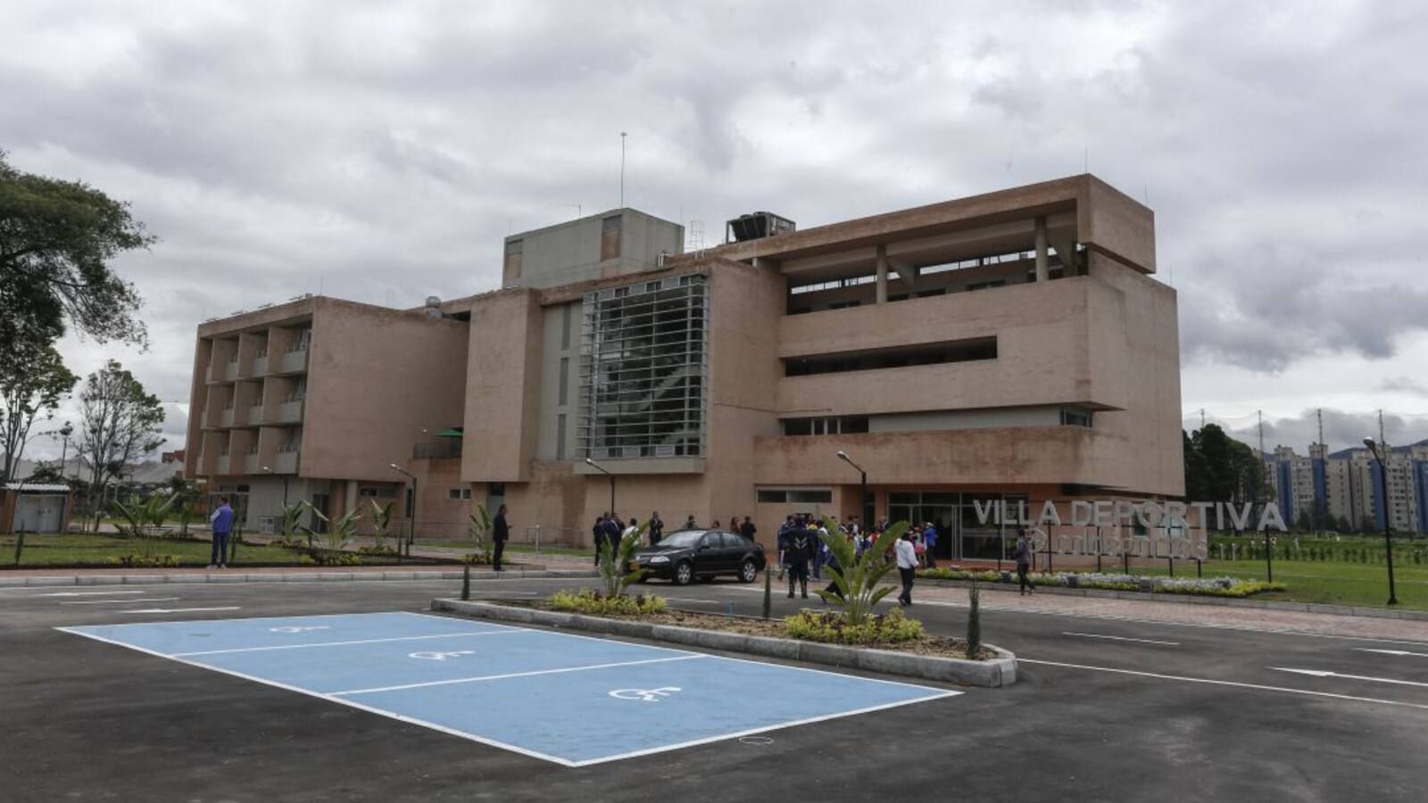 Edificio moderno de color beige con vidrio, cielo nublado, gente cerca de la entrada, lugares de estacionamiento azules.