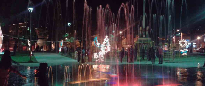 Exhibición de fuentes por la noche con chorros de agua de colores, árbol de Navidad y gente en una plaza de la ciudad iluminada.