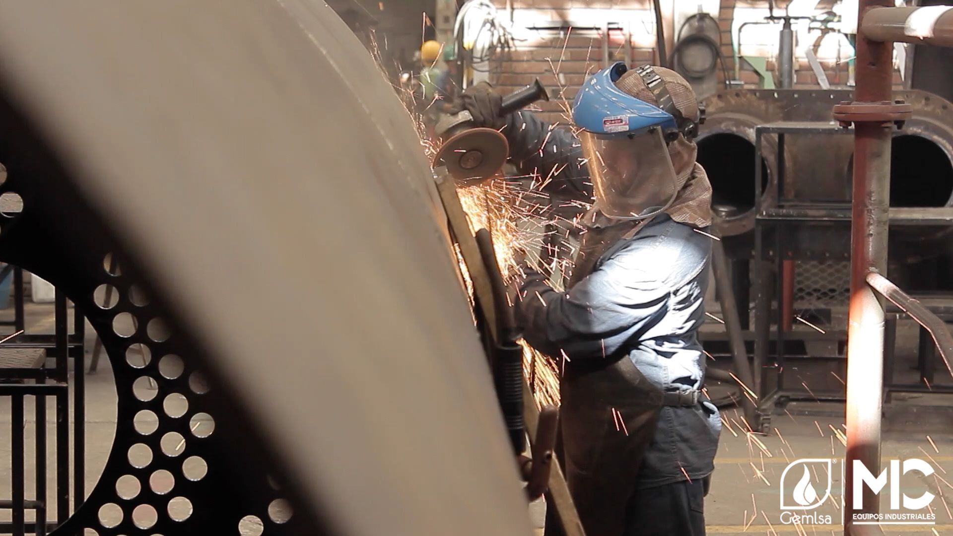 Una persona con equipo de protección muele una gran estructura metálica mientras saltan chispas en el taller.