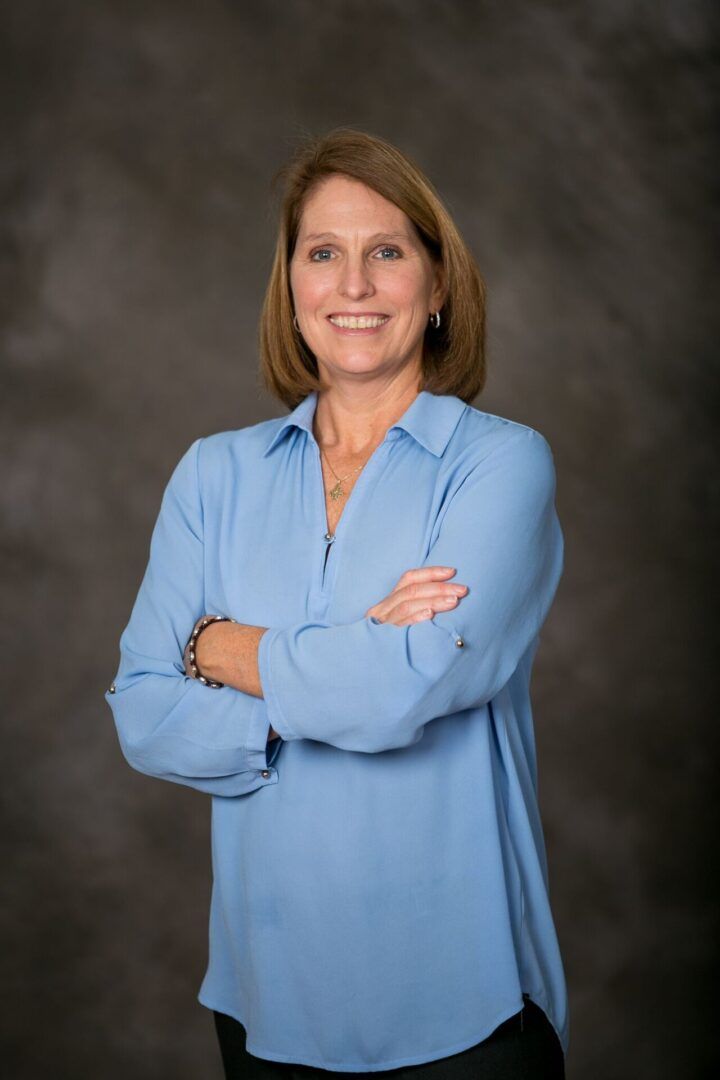 A woman in a blue shirt is standing with her arms crossed and smiling.
