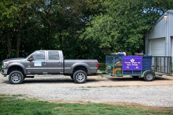 Silver pickup truck towing a purple utility trailer in a gravel yard beside a metal building