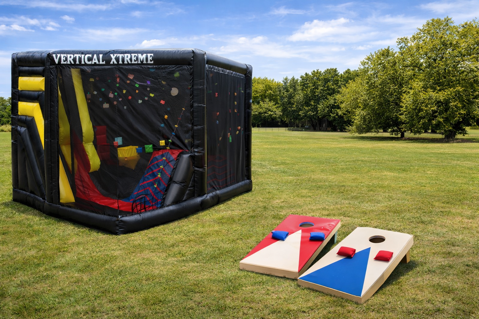 Inflatable rock climbing wall with cornhole boards on grass under blue sky.