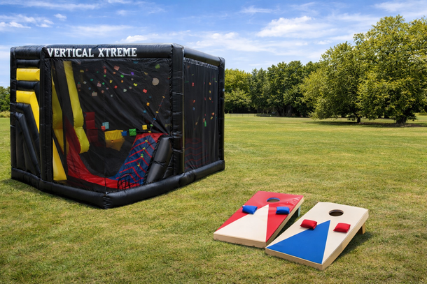 Inflatable rock climbing wall with cornhole boards on grass under blue sky.