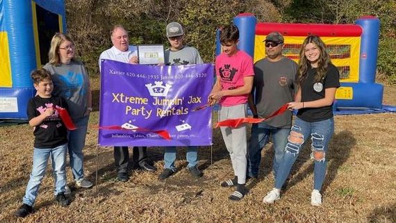 Group holding a purple Xtreme Jumpin Jax Party Rental banner in front of a colorful inflatable bounce house.