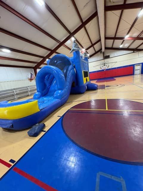 Blue inflatable obstacle course in a gymnasium with a yellow slide and red, blue, and tan floor markings