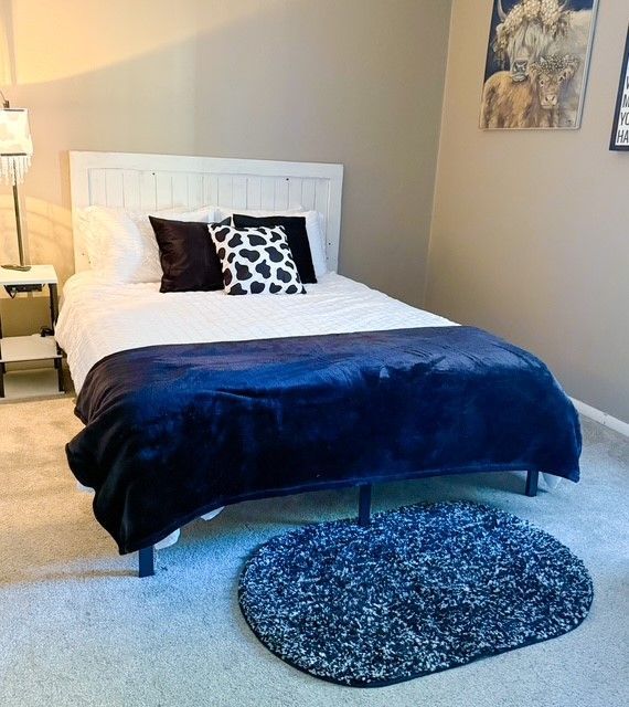 Bedroom with a white headboard, dark blue blanket, pillows, and a round rug.