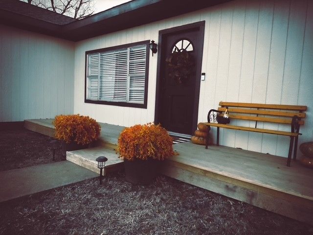 Front porch with brown door, window with blinds, bench, orange bushes, and door wreath.