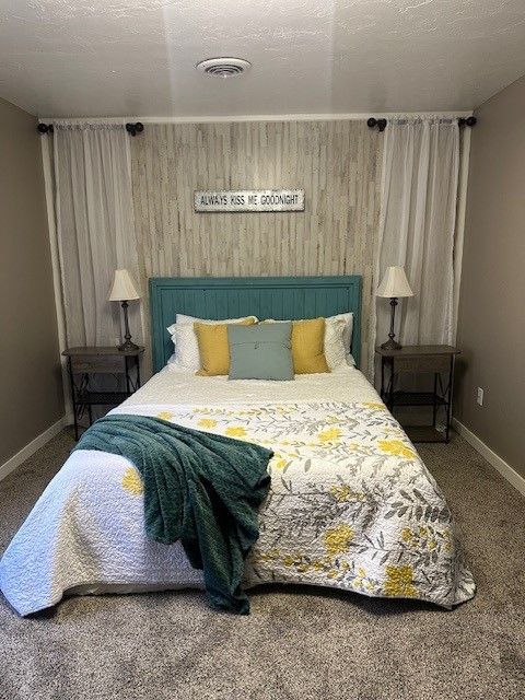 Bedroom with teal headboard, floral quilt, yellow pillows, side tables, and sheer curtains.