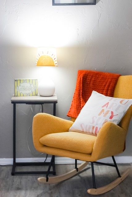 Yellow rocking chair with orange throw and pillow, next to a table with a lamp and wall art.