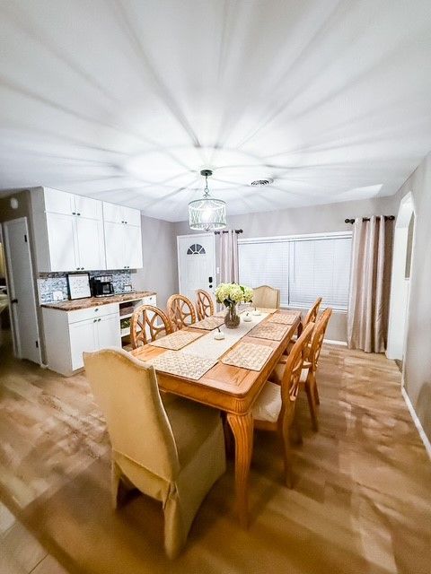 Dining room with a long wooden table, chairs, and white cabinets. Light streams in from a window.