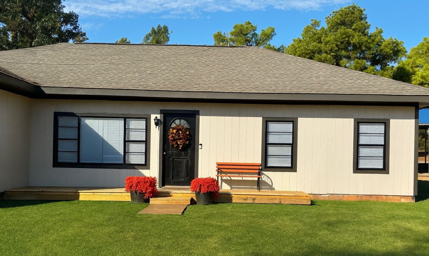 Single-story beige house with black shutters, front porch, and red flowers on a sunny lawn