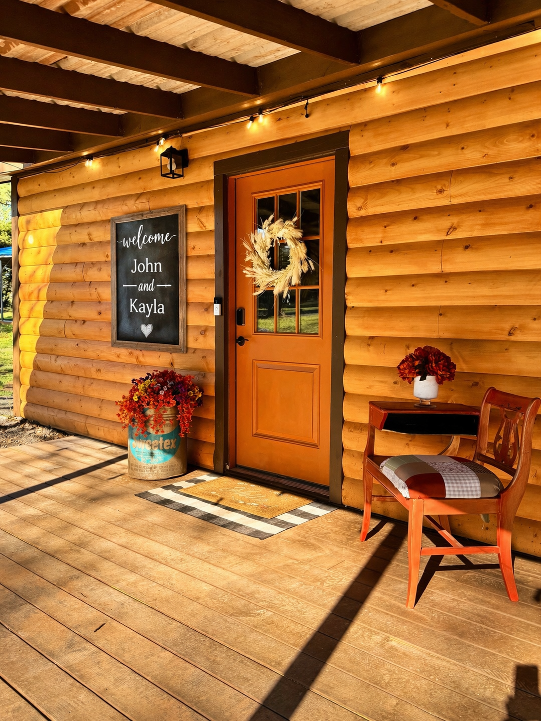 Sunny wooden porch with orange door, chalkboard sign, wreath, and small table with flowers and chair
