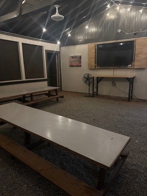 Indoor space with long tables, benches, and a mounted TV. String lights hang from the ceiling.