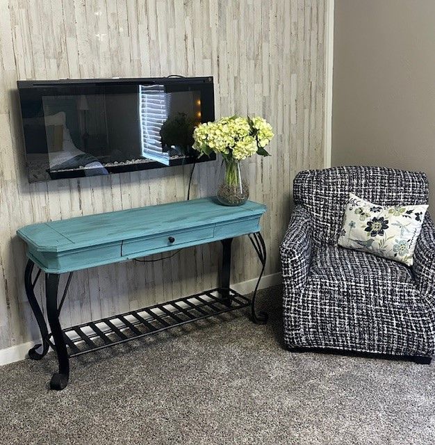 A blue console table with black metal legs, under a wall-mounted fireplace and next to a patterned armchair.