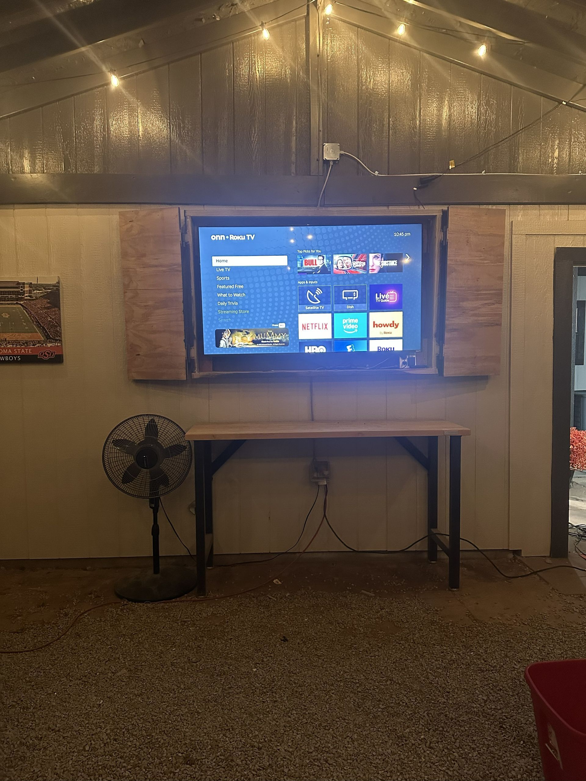 TV mounted on wooden planks, above a table. A fan sits left. The space has string lights and a gravel floor.