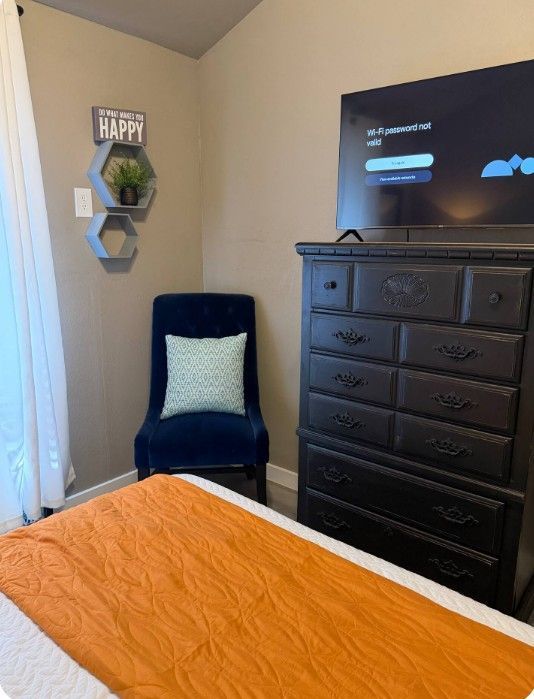 Bedroom corner with blue chair, black dresser, wall decor, and TV; orange bedspread in foreground