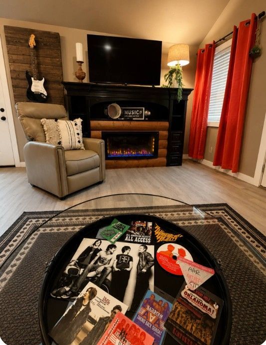 Cozy living room with fireplace, TV, armchair, red curtains, and a patterned rug with magazines on the floor