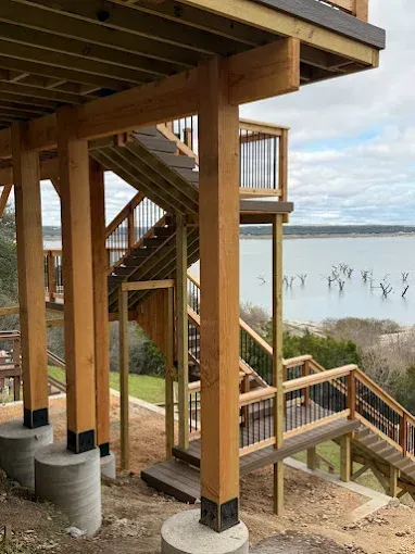 Wooden stairways and deck posts on a lakeside home overlooking calm water and a grassy shore