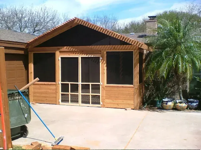 Front view of a small wooden house with a gabled roof and concrete driveway, flanked by a palm tree.