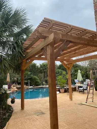 Wooden pergola beside a backyard pool with patio chairs and palm trees