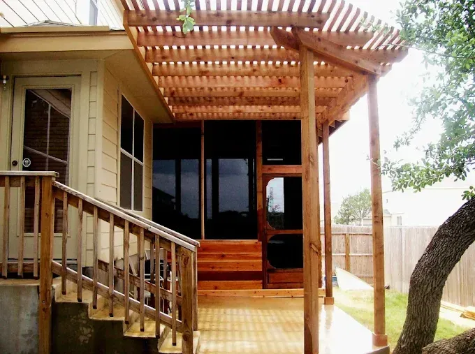 Wooden porch with pergola roof and steps beside a beige house, surrounded by trees.