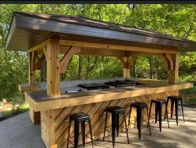 Covered outdoor bar with stools and a built-in grill under a wooden pavilion in a wooded setting